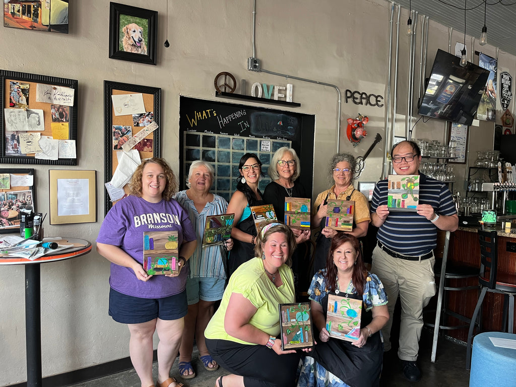 A group of nine people posing together in a cafe or workshop setting, each holding a colorful square art print or book with a landscape illustration. The background features a chalkboard, framed pictures, and bar-style decor.