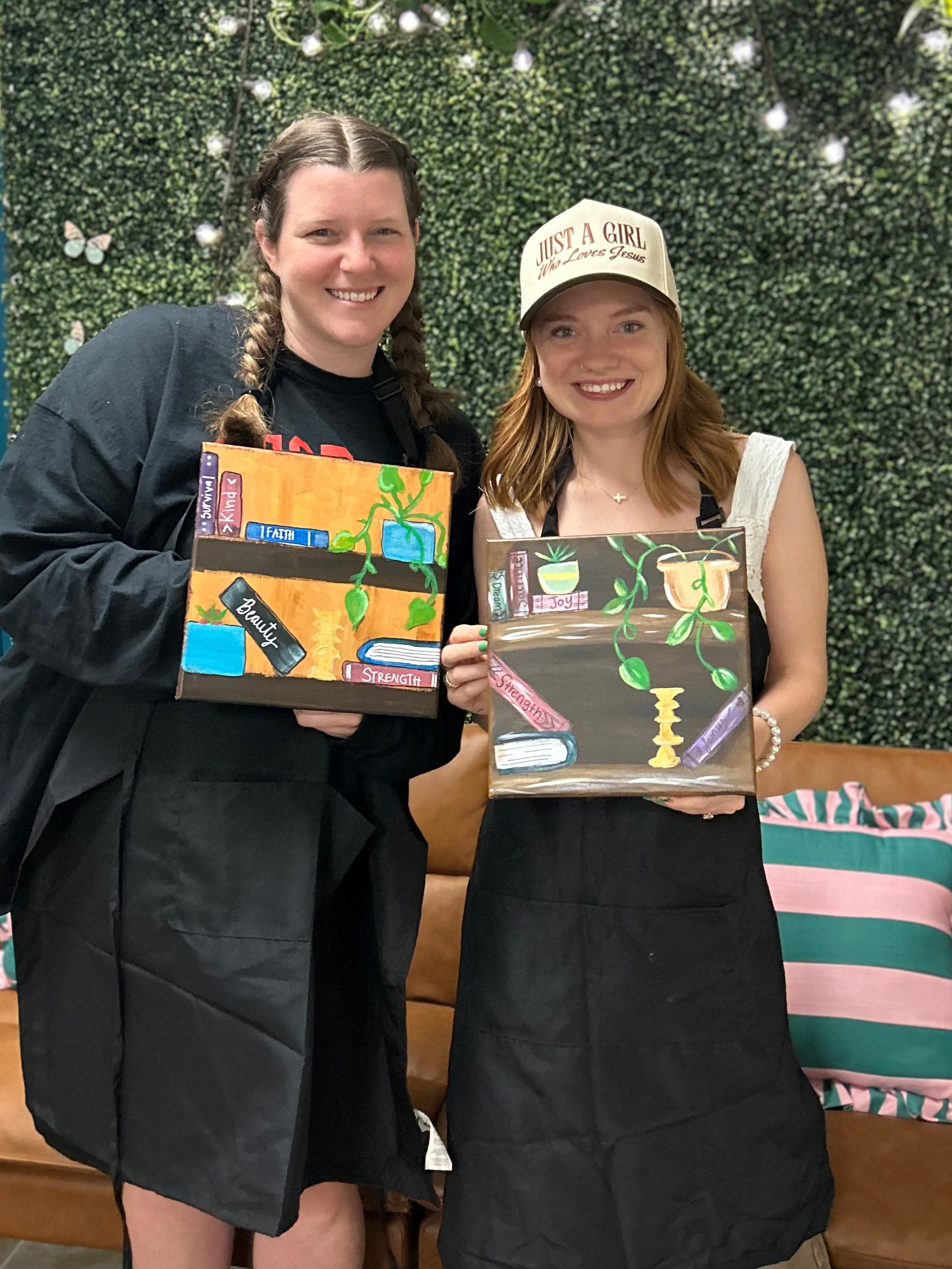Two women holding paintings in a casual indoor setting with a green wall and couch.