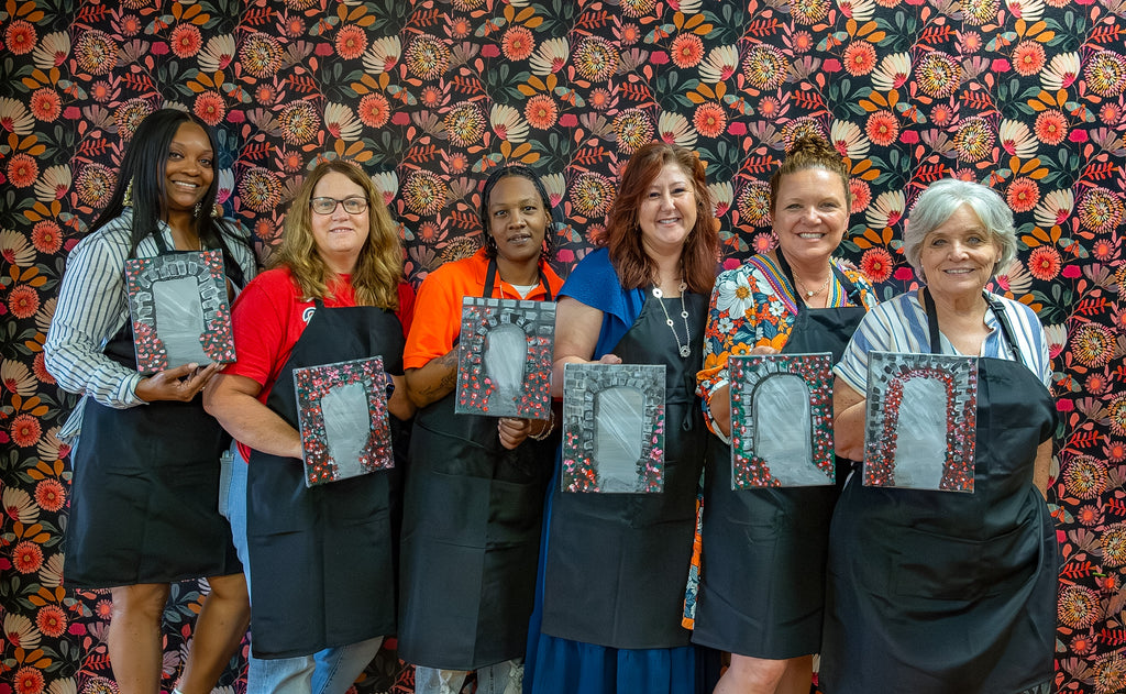 Group of women wearing aprons holding framed artworks against a floral patterned wall.